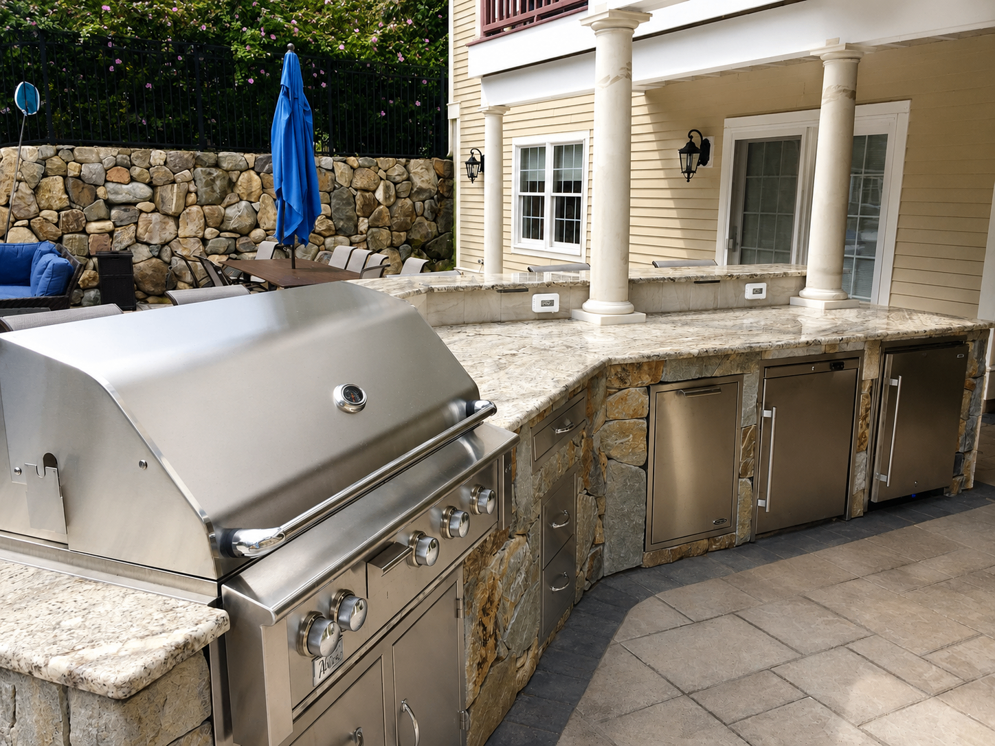 Granite outdoor kitchen with stainless steel grill, refrigerator and stone base beside a covered patio