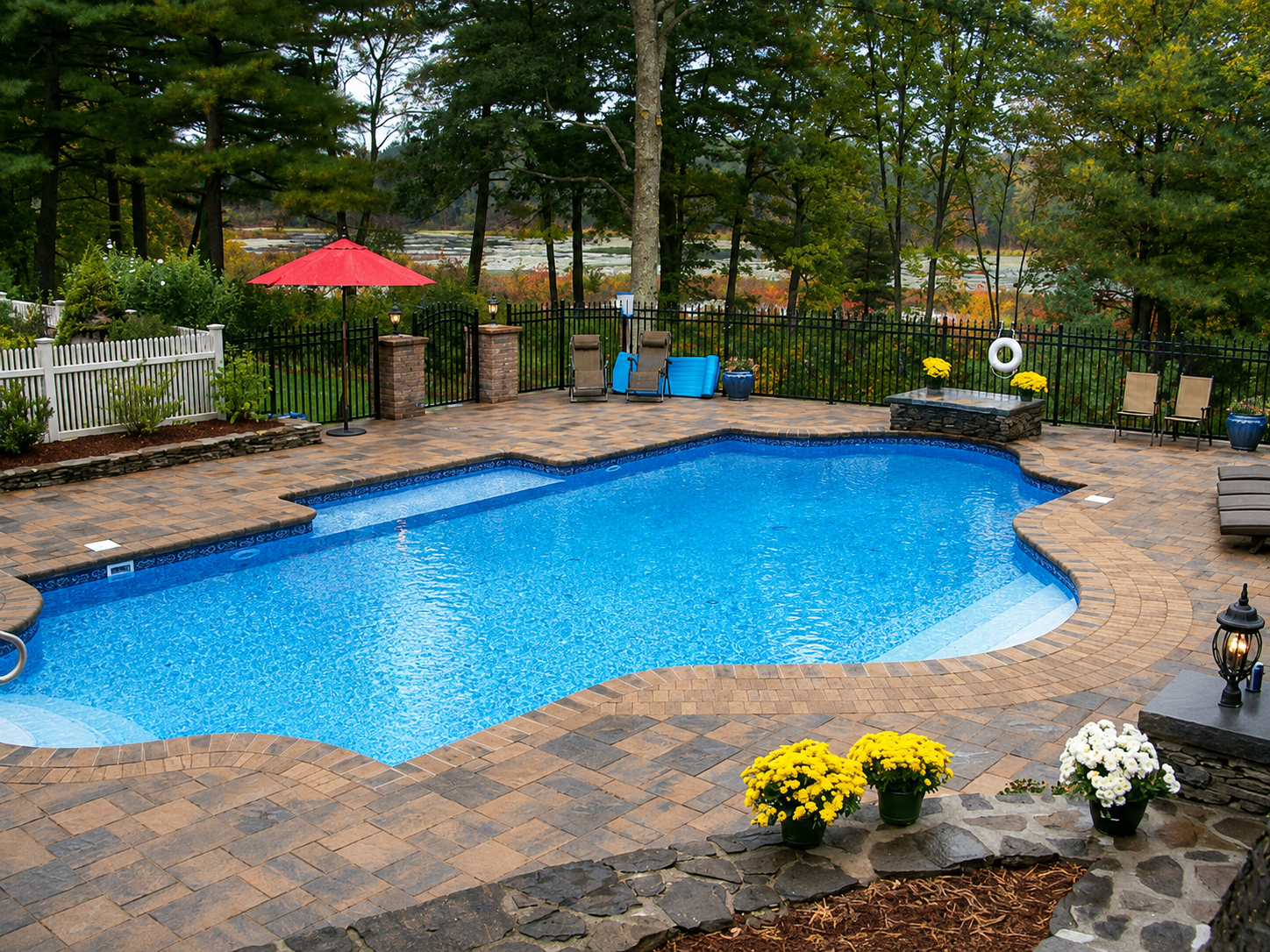 Paver pool deck surrounding a blue swimming pool with stone borders and autumn trees in the background